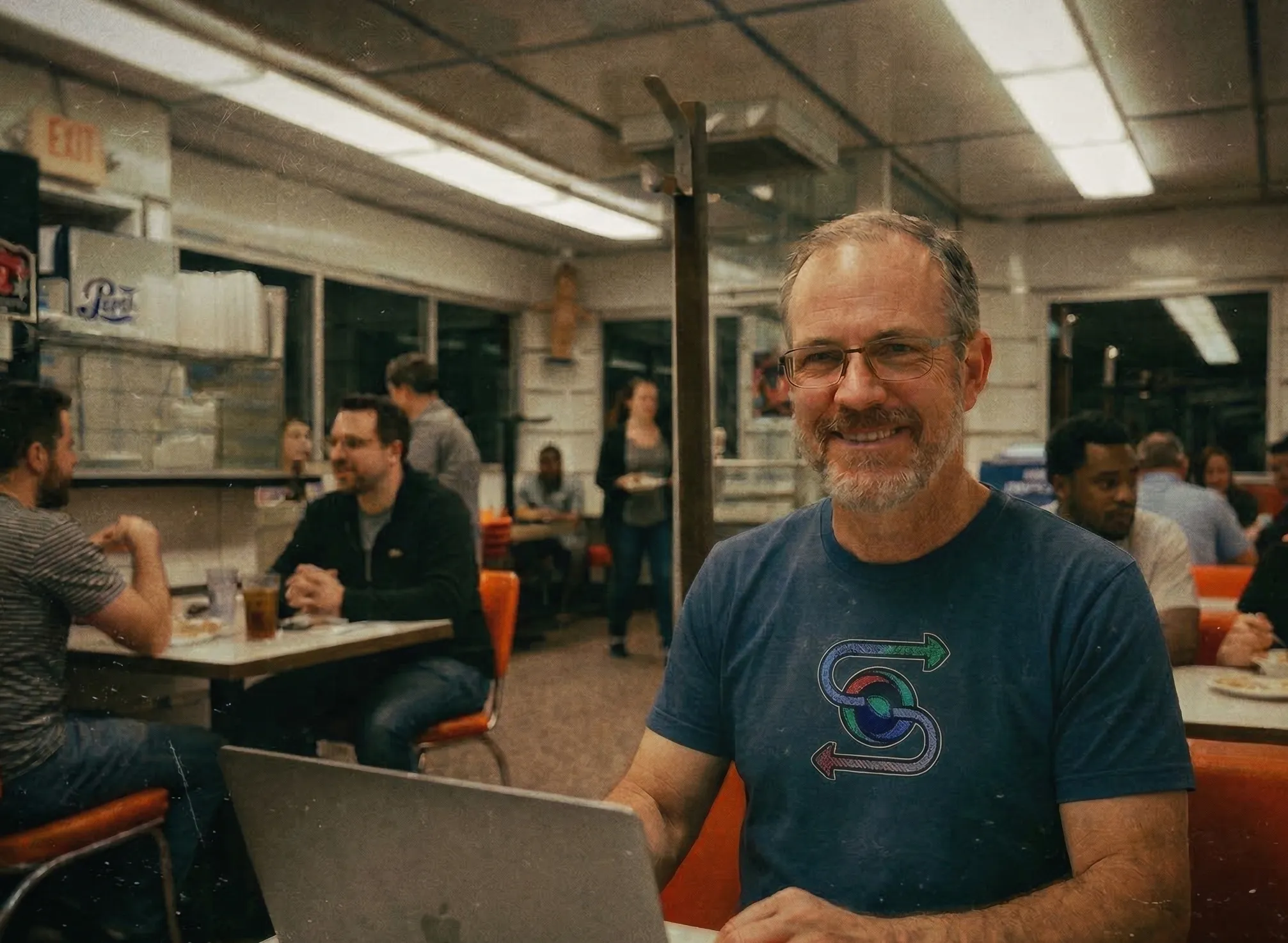 Ned Ludd smiling at his laptop in a busy diner, wearing glasses and a blue t-shirt